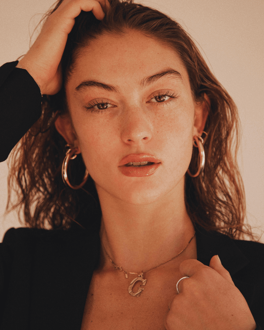 A woman with wavy hair poses, wearing recycled gold hoop earrings and a sustainable gold initial necklace