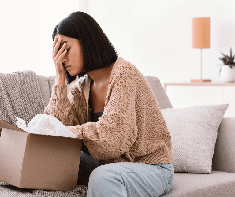 A lady covers her face with her hand as she sits in front of a delivery box.